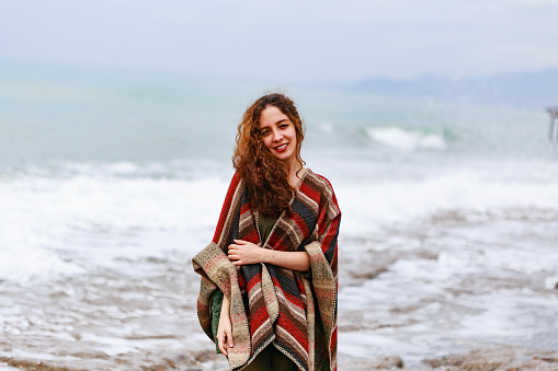 Portrait of happy brunette woman by seaside wearing striped red black and beige poncho