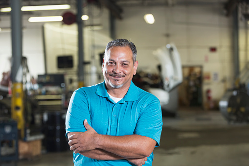 A mature, Hispanic man in his 40s, standing with his arms folded, smiling confidently at the camera, in a vehicle repair shop. In the background out of focus is a truck cab with an open hood.
