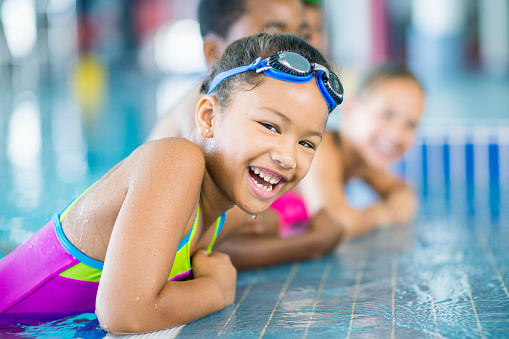 A multi-ethnic group of children are at a swimming pool. They are wearing swimsuits. A girl wearing goggles is taking a break from swimming, and smiling at the camera.