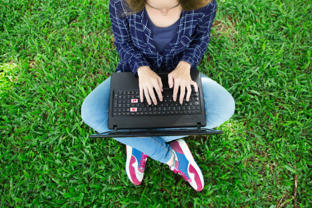 Cropped view of young woman working on laptop computer and sitting on grass in park. Top view.