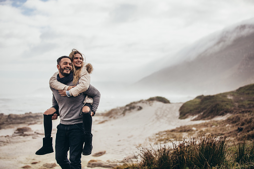 Man carrying girlfriend on his back at winter beach. Young couple enjoying holidays at the seashore.