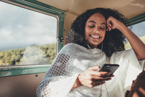Woman enjoying on her road trip African woman on road trip travelling by van. Smiling female sitting on backseat of car and  listening music with mobile phone. back seat photos stock pictures, royalty-free photos & images