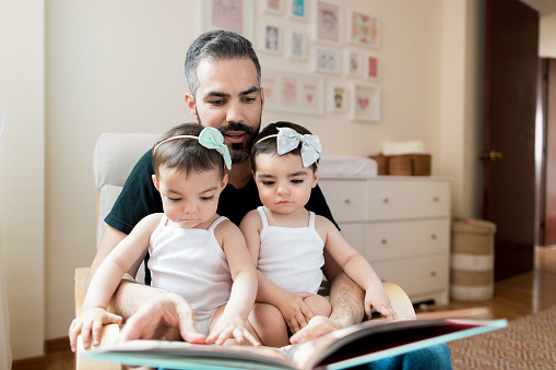 Latin father reading book to baby twins on lap