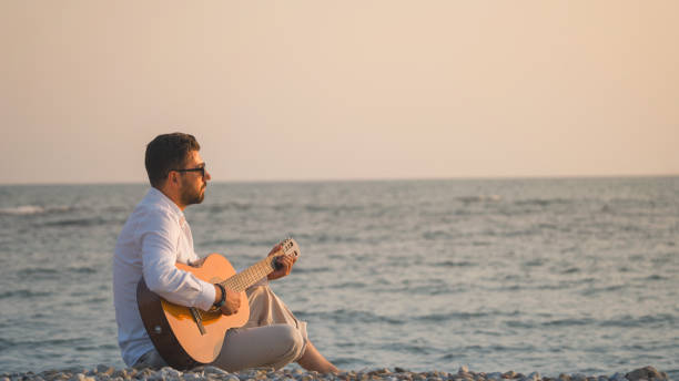 Young attractive brown boy dressed in summer form and a hat walking on the shore of a mediterranean beach holding a Spanish guitar