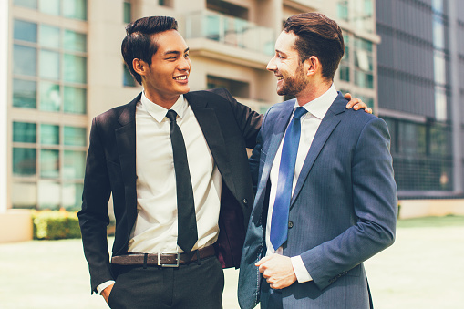 Closeup of two smiling business men looking at each other, chatting and embracing with office building in background
