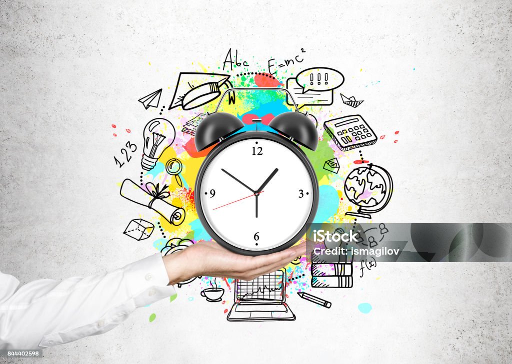 Man s hand and a time management scheme Close up of a hand of an unrecognizable businessman holding an alarm clock with a time management sketch around it near a concrete wall. Time Stock Photo