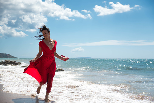 Beautiful young woman in long red dress with long black hair, walking on a yellow sandy beach by the ocean and looking down. Shot made in Greece, Corfu island in September 2017.