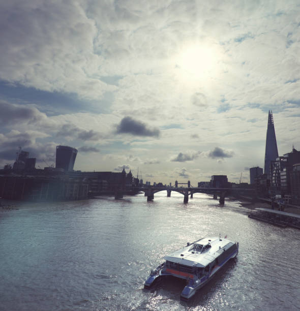 Sun shining through the clouds on skyscrapers of London city over river Thames with passenger boat on the foreground. Counterlight toned panoramic image.