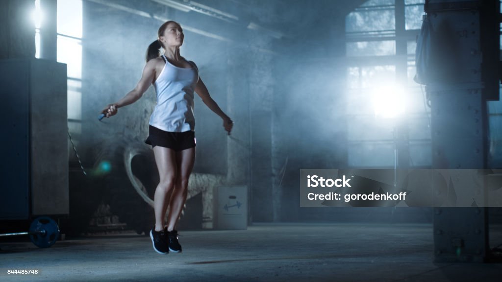 Athletic Beautiful Woman Exercises with Jump / Skipping Rope in a Gym. She's Covered in Sweat from Her Intense Cross Fitness Training. Dark atmosphere. Gym Stock Photo