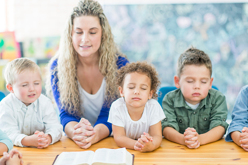 A multi-ethnic group of children are indoors during Sunday school class. They are wearing regular clothing. They are sitting at a table and praying with their teacher.