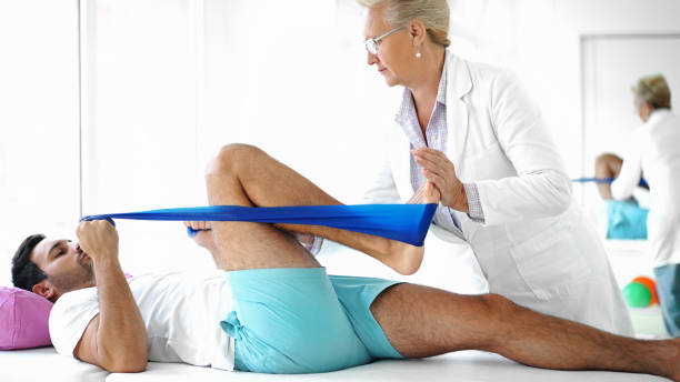 Young man in physical therapy. Closeup side view of late 50's female doctor assisting an early 30's male athlete during physical therapy treatment. The patient is laying on back and doing leg presses each leg at a time and making it harder with a resistance band. physical therapy stretching stock pictures, royalty-free photos & images