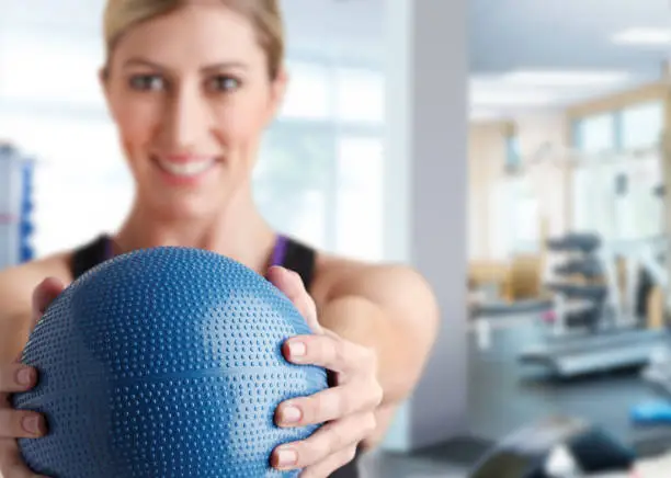 A young female patient holds a blue medicine ball as she looks at and smiles directly at the camera while she exercises in a physical therapy clinic.  The image is photographed with a very shallow depth of field with the focus on the ball in the foreground.