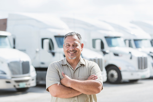 An Hispanic man in his 40s standing in front of a row of parked semi-trucks outside a distribution warehouse.