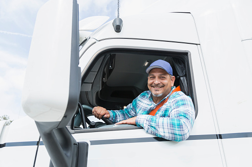 A mature Hispanic man in his 40s behind the wheel, driving a semi-truck. He is looking at the camera, smiling through the open driver's side window.