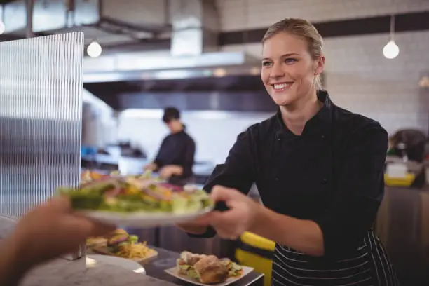 Attractive young female chef giving fresh Greek salad to waiter at coffee shop