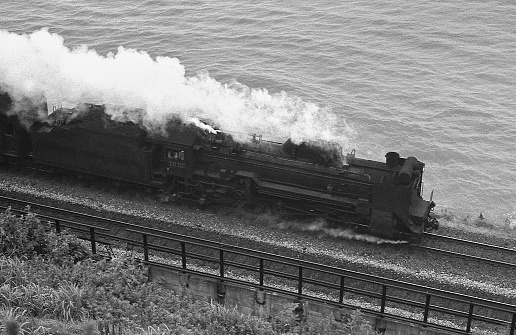 Steam train driving in the countryside with smoke coming from the chimney. The black and red locomotive is pulling passenger railroad car with tourists.