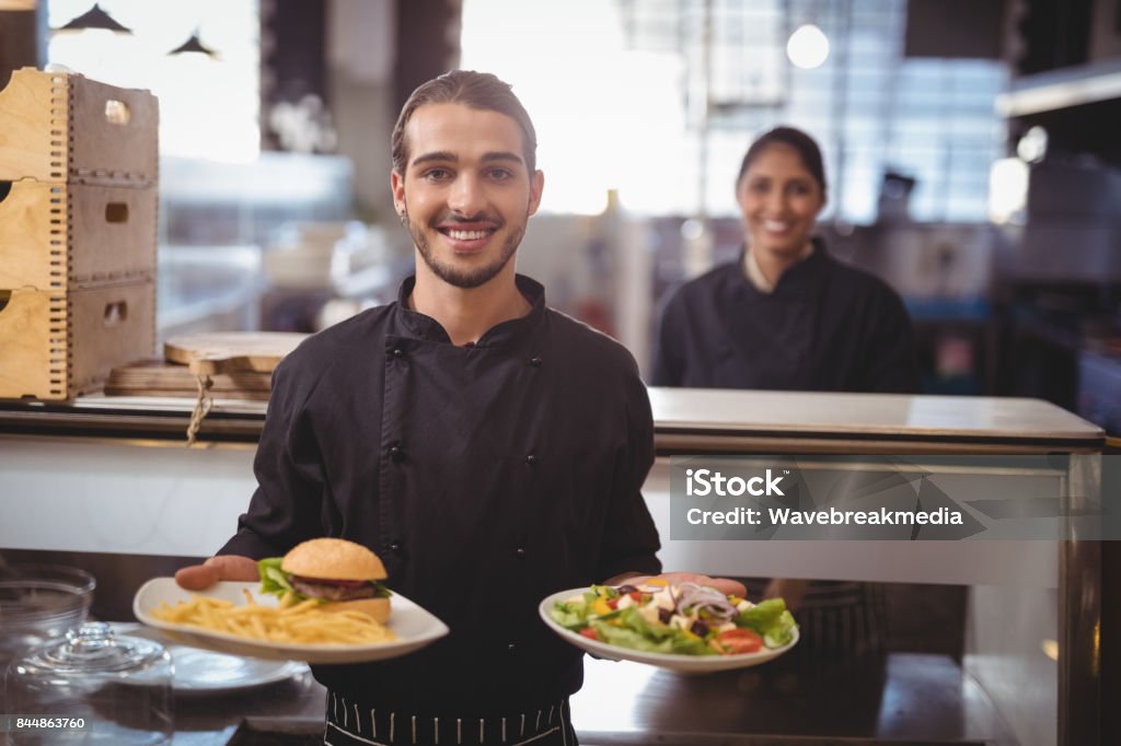 Portrait of smiling young waiter serving food while standing against waitress Portrait of smiling young waiter serving food while standing against waitress at counter in coffee shop Serving Food and Drinks Stock Photo