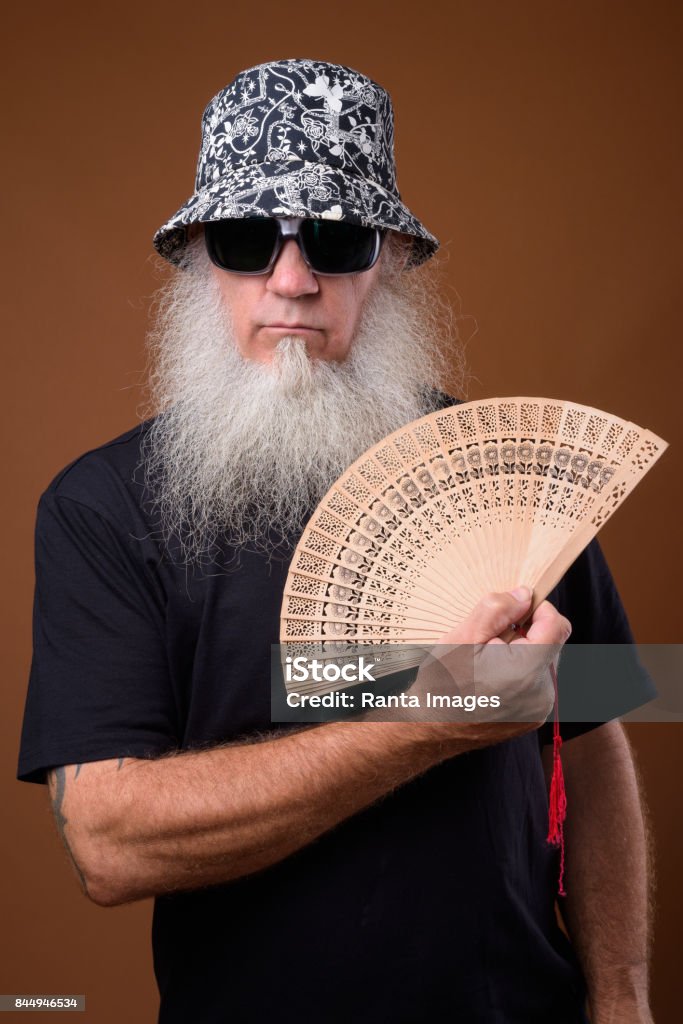 Portrait of bald man with long gray beard holding hand fan Studio shot of bald man with long gray beard holding hand fan while wearing bucket hat and sunglasses against colored background vertical shot Hand Fan Stock Photo