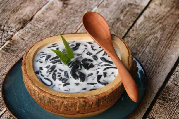 Es Dawet Ireng, the Javanese cold dessert of black cendol (tapioca short-noodle jelly) made from toasted paddy straw. It is served with crushed ice, coconut milk, and palm sugar syrup in a thick wooden bowl that has been carved out of single wooden block. A ceramic plate of solid color underlines the bowl. A wooden spoon is placed on the bowl rim. The dessert is arranged on a rustic wooden table.