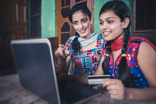 Indoor image of happy sisters doing online shopping through internet by using credit/debit card on laptop at their traditional village house. They are in their traditional dress of north India, (salwar kameez and dupatta). Two people, waist up, selective focus with copy space.