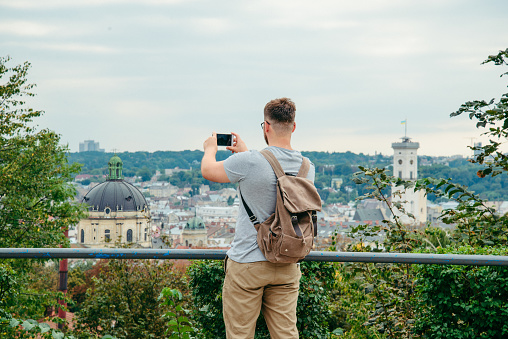 man looking at old european city from the top of the hill and taking a picture on his phone