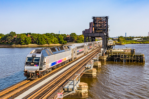 Rutherford, United States – August 9, 2017: Bombardier NJ Transit commuter train with state-of-the-art dual-powered Bombardier ALP-45DP locomotive #4504 and bilevel (Multi-level, double-decker) Bombardier stainless steel cars crossing the HX Drawbridge over the Hackensack River enroute to Suffern.  In the background is the World Trade Center tower. The bascule bridge built in 1911 carries both the Bergen County and Pascack Valley lines.  Horizontal, copy space.