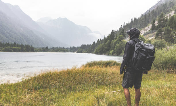 wanderer mit einem rucksack beobachten den see umgeben von bergen - regenmantel stock-fotos und bilder