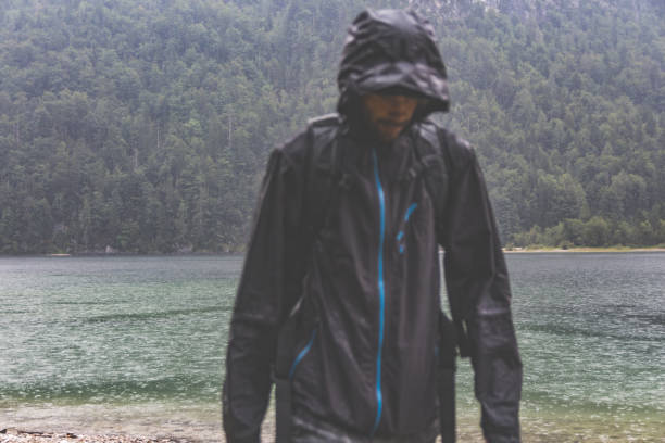 istock Man wearing a rainjacket walking towards the camera in heavy rain with mountains, forest and a lake in the background 845235586