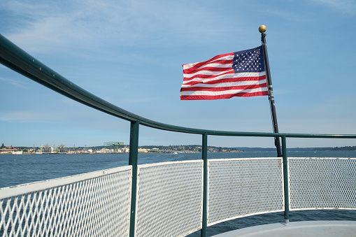 Amaerican flag blowing in breeze at stern of passenger ferry crossing Sinclair Inlet from Bremerton, Washington to Port Orchard, Washington with Puget Sound Naval Shipyard in background
