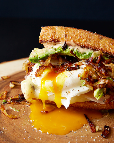 Poached egg sandwich with caramelized onion, avocado, meat and cheese on a wooden cutting board