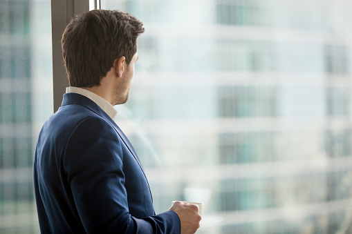 Businessman in blue business suit standing with coffee cup in hand looking at skyscraper. Company leader taking break during work day, dreaming of success, considering investment decision. Back view