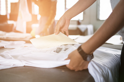 Young woman pull out paper from waterproof film on fabric. worker working on manual screen printing on t-shirt at her shop.