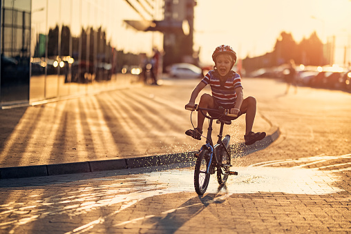 Little boy riding a bike in urban area. The boy is shouting and riding a bike through a puddle. Splashing water backlit by the setting sun.\n