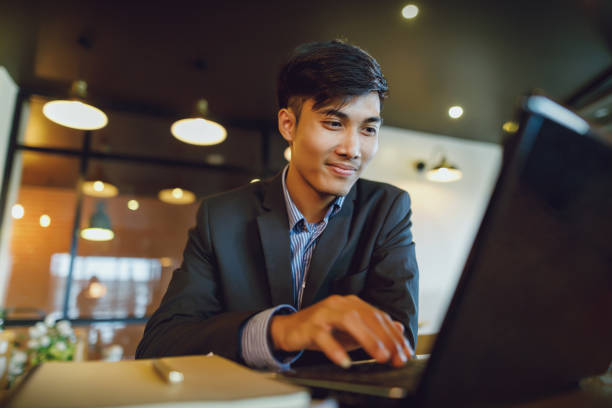 lachende aziatische zakenman in pak werken met laptop - male employee office stockfoto's en -beelden