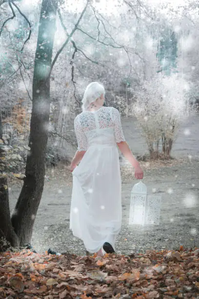 A woman goes with a white dress and lantern through a forest with snowfall