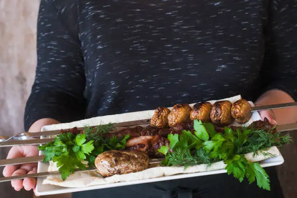 Waiter brings shish-kebab with grilled vegetables on white dish. Meat, field mushrooms and fresh green herbs, barbecue and natural food preparing in restaurant