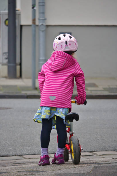 Little girl with bike helmet and balance bike waiting at the curb to cross the street stock photo