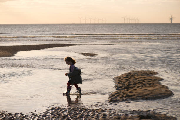 little girl with offshore windfarm a little girl walks across a windy beach splashing in the sea , with offshore wind farms in the distance liverpool one stock pictures, royalty-free photos & images