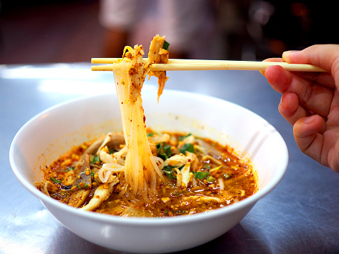 Thai local traditional spicy chicken piece with thin white rice noodle soup, gripping up with wooden bamboo chopsticks by right hand, served in white plastic bowl, on stainless steel table