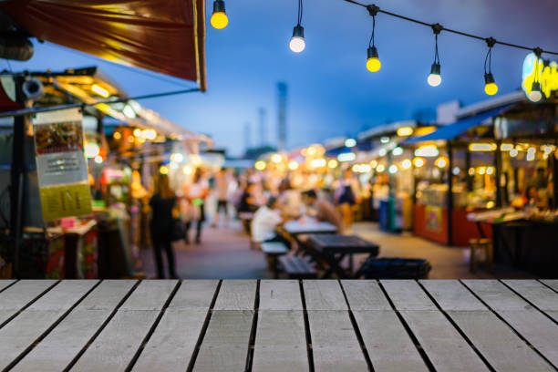 immagine di tavolo in legno di fronte alla lampadina decorativa a luci a corda all'aperto nel mercato notturno con persone sfocate, festival e concetti di vacanza, può essere utilizzata per esporre o montare i tuoi prodotti. - outdoors market foto e immagini stock