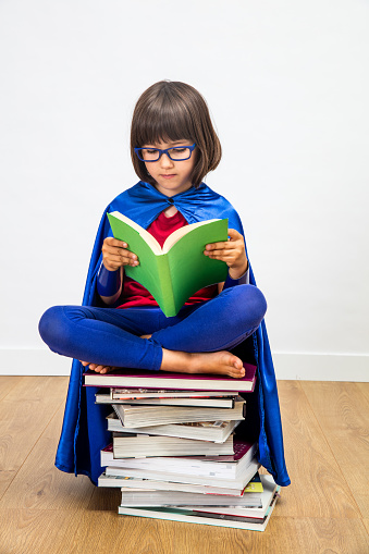 wise 6-year old schoolgirl with a super hero costume reading seated on top of a stack of books reading with eyeglasses for girl power in education and female knowledge, indoor concept