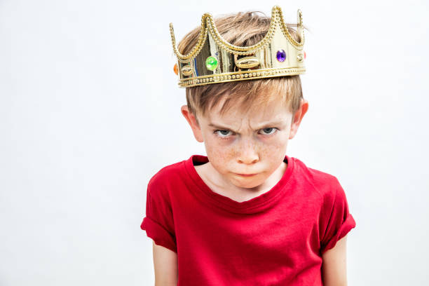 irritated beautiful spoiled boy with frowning freckles and a dirty look wearing a golden crown for mad attitude facing parenthood and education, white background, indoors