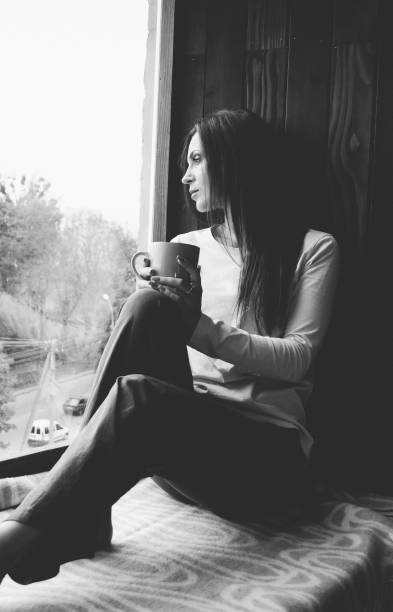 istock Beautiful young woman drinking coffee and looking through window while sitting at windowsill at home 845897710