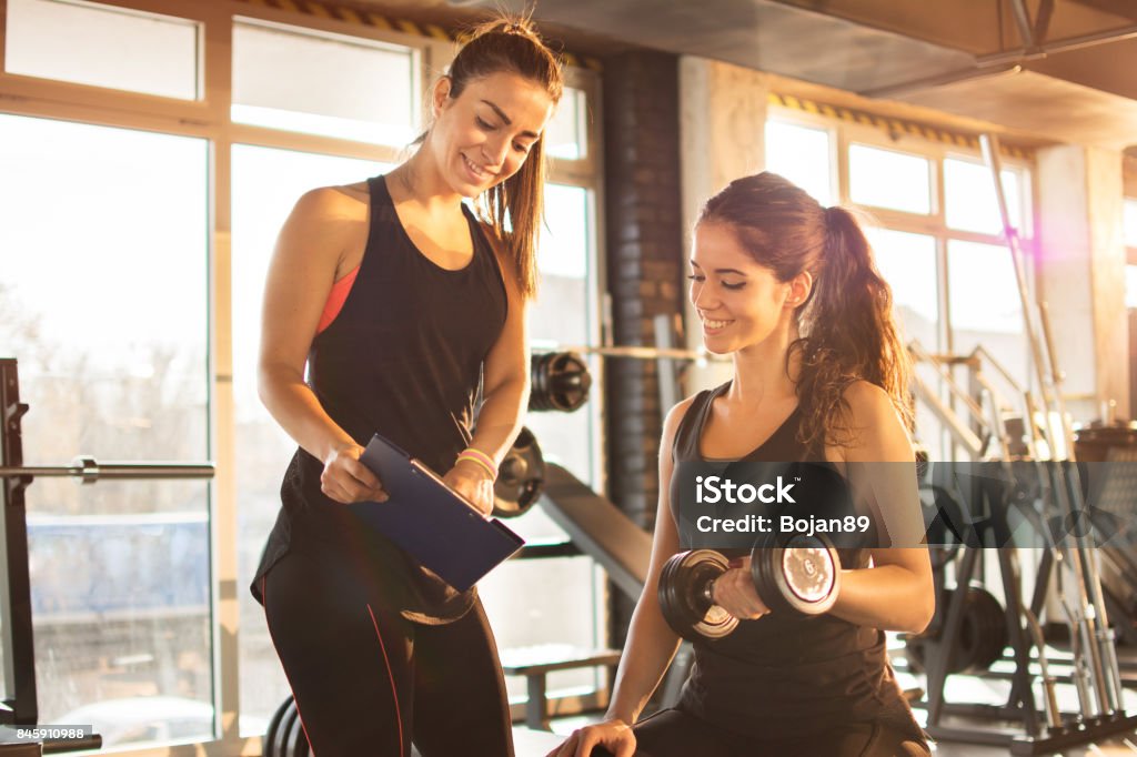 Female fitness instructor showing exercise progress on clipboard to young athletic woman at gym. Fitness Instructor Stock Photo