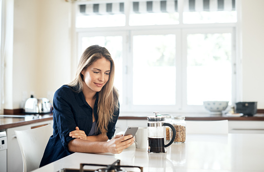 Shot of a young woman using a mobile phone while having breakfast in the kitchen at home