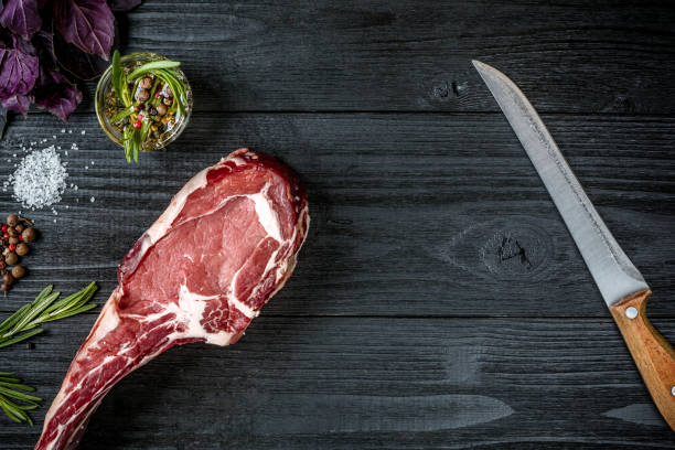 Fresh raw beef with basil and a sprig of rosemary with knife with a wooden handle on black wooden background. Top view