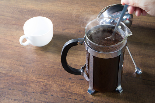 Coffee brewing preparation with french press coffee plunger mug as brewing instrument with a spoon stirring in motion blur and steam over the hot drink surface. Presented in warm tone.
