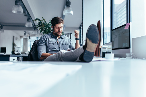 Young man listening music on phone at the workplace with feet on the table. Businessman relaxing in office during break.