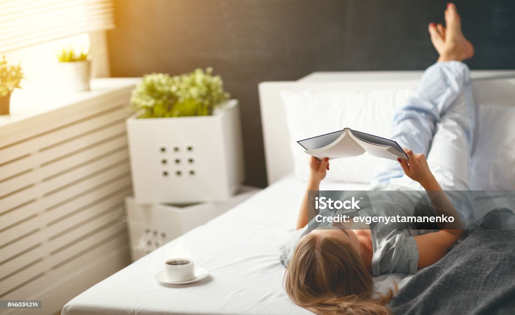 Happy young woman reads  book and drinks coffee in bed Happy young woman reads book and drinks coffee in bed in   morning Relaxation Stock Photo