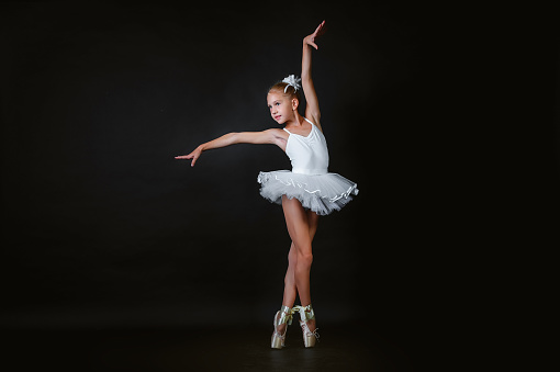 A small young ballerina performs an element of ballet dance on a black background.